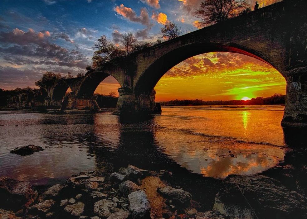 Dramatic sunset viewed through the main arch of the Interurban Bridge, golden light reflecting on the Maumee River