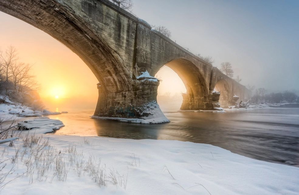 The Interurban Bridge at sunrise in winter, arches framing the rising sun over a frozen Maumee River
